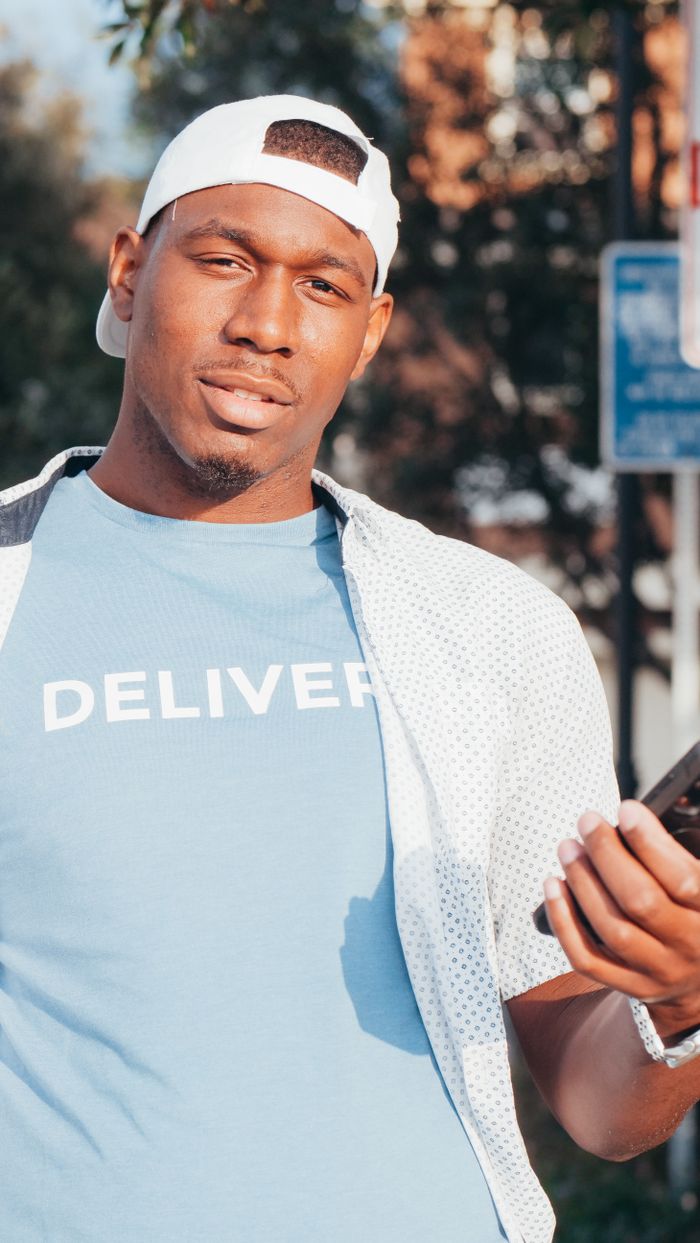About Casual delivery person holding a smartphone outdoors, wearing a 'Deliver' shirt and cap.